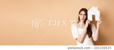 Real estate. Upset young woman showing paper house cutout and frowning sad, looking aside thoughtful, thinking of buying apartment, standing on beige background Real estate. Upset young woman showing paper house cutout and frowning sad, looking aside thoughtful, thinking of buying apartment, standing on beige background 103150525
