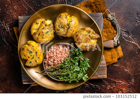 Crushed, smashed potatoes baked with rosemary and thyme on plate. Dark background. Top view Crushed, smashed potatoes baked with rosemary and thyme on plate. Dark background. Top view 103152512
