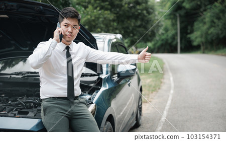 Asian businessman stranded on roadside after car breakdown, using phone to call for help. Candid shot of man waiting for assistance and thumbing up. Transportation and technology concept. Asian businessman stranded on roadside after car breakdown, using phone to call for help. Candid shot of man waiting for assistance and thumbing up. Transportation and technology concept. 103154371
