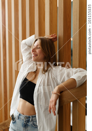 Close up portrait caucasian young happy woman with fresh and clean skin stands next to wooden decorated wall. Wear white shirt. Lifestyle, relax, female beauty concept Close up portrait caucasian young happy woman with fresh and clean skin stands next to wooden decorated wall. Wear white shirt. Lifestyle, relax, female beauty concept 103155835