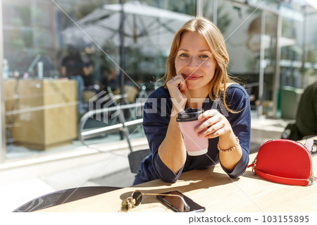 A pretty young hipster girl with cup of drink is sitting at a table of a street summer cafe.Take a coffee brake 103155895