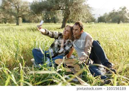 Couple taking selfie or making video call while spending time together on picnic with guitar in green summer grass Couple taking selfie or making video call while spending time together on picnic with guitar in green summer grass 103156189