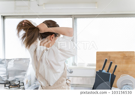 Young woman tying her hair up before starting housework in the kitchen 103156530