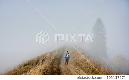 Man hiker strolling down hiking path on grassy hill with misty trees and sky in fog on background 103157271