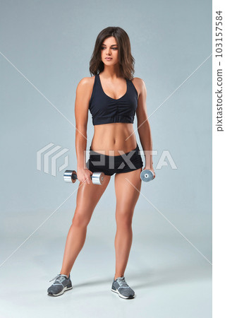 Athletic woman pumping up muscles with dumbbells on gray background 103157584