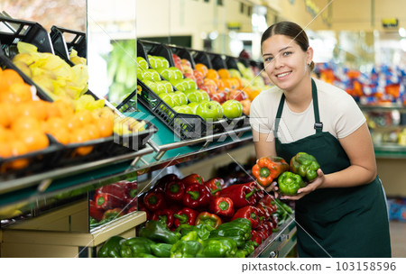 Positive girl seller in an apron offers fresh bell peppers 103158596