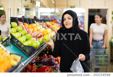 Asian girl in hijab buys ripe apples in supermarket Asian girl in hijab buys ripe apples in supermarket 103158599