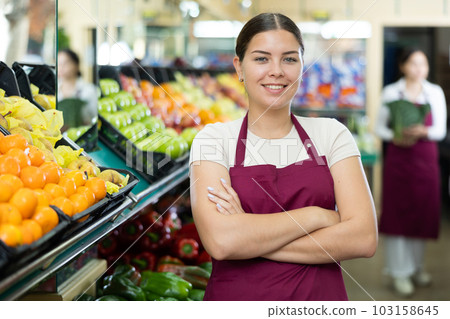 Smiling woman seller wearing apron standing near fresh tangerinas on supermarket 103158645