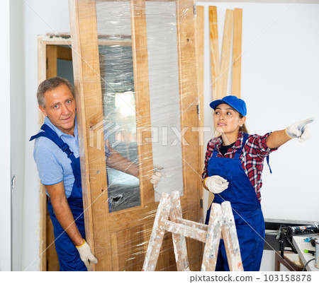Female carpenter pointing with the hand while a repairman in uniform dragging the door construction into an apartment 103158878