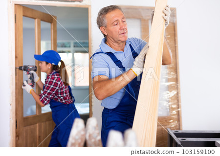 Confident middle-aged repairman in uniform checking wooden timber at a construction site in an apartment Confident middle-aged repairman in uniform checking wooden timber at a construction site in an apartment 103159100