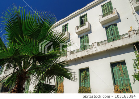 A low-rise deserted 1930s hotel with a decayed facade and green wooden window shutters under the summer blue sky. 103165384