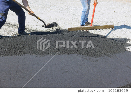 Work laying asphalt on road pavement is accompanied by men shovelling black gravel for purpose of laying asphalt 103165639