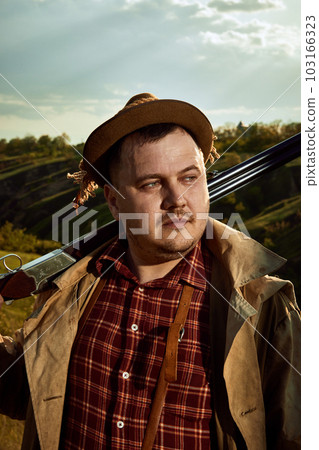 Close up portrait with handsome hunter wearing checked shirt and hat holding weapon on shoulder after hunting over nature background 103166323