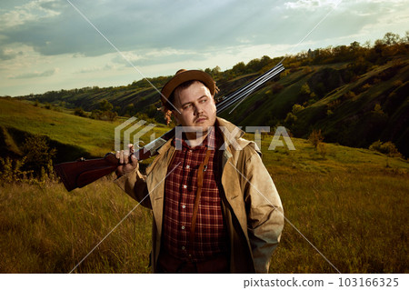 Hunter wearing vintage style checked shirt and coat holding shotgun on shoulder after hunting and looking away over nature background 103166325