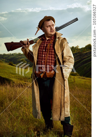 Portrait with experienced hunter wearing checked shirt and hat holding rifle on shoulder and resting after hunting over nature background 103166327