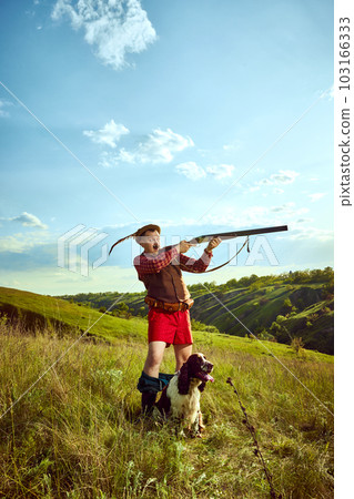 Portrait with funny hunter wearing retro hat with feather holding gun and shooting with dog English springer spaniel over nature landscape background 103166333