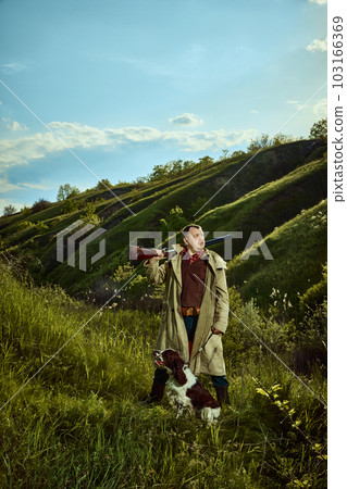 Male hunter wearing retro coat holding gun and standing with companion dog English springer spaniel over nature landscape background. Spring hunting season 103166369