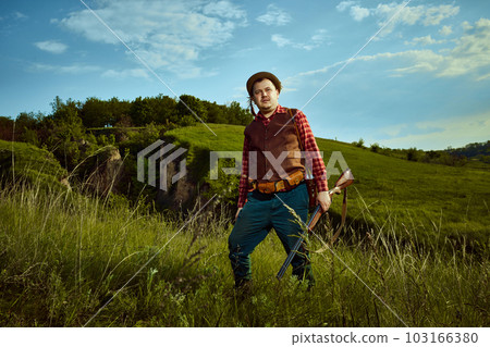 Portrait with adult man, hunter wearing vintage clothes holding weapon in hand and looking at camera over wonderful nature background 103166380
