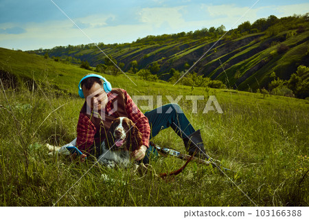 Portrait with man, hunter sitting on grass with his dog English springer spaniel listening to music and resting after hunting on nature landscape background 103166388
