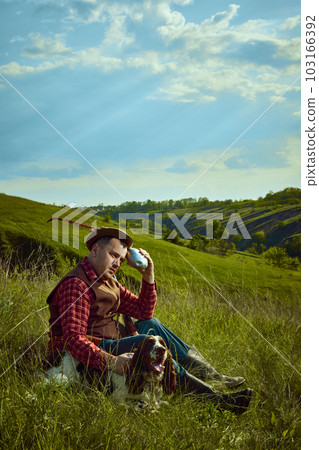 Portrait with man in vintage clothes sitting on grass with his dog English springer spaniel and resting after hunting on nature landscape background. Hot summer day Portrait with man in vintage clothes sitting on grass with his dog English springer spaniel and resting after hunting on nature landscape background. Hot summer day 103166392