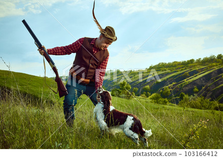 Portrait with man, vintage hunter wearing retro hat with feather holding weapon and petting the dog English springer spaniel over beautiful nature landscape background Portrait with man, vintage hunter wearing retro hat with feather holding weapon and petting the dog English springer spaniel over beautiful nature landscape background 103166412