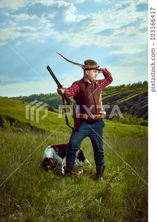 Portrait with male vintage hunter holding weapon standing with companion dog English springer spaniel and seeking for prey over nature landscape background Portrait with male vintage hunter holding weapon standing with companion dog English springer spaniel and seeking for prey over nature landscape background 103166417
