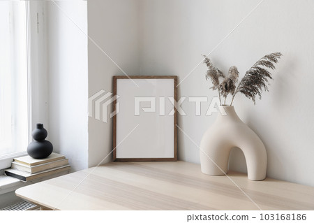Vertical wooden picture frame, poster mockup in the corner. Wooden table, desk. Modern organic shaped vase. Dry flowers, grass. Old books on window sill. Home staging. Minimal Scandi boho interior. 103168186