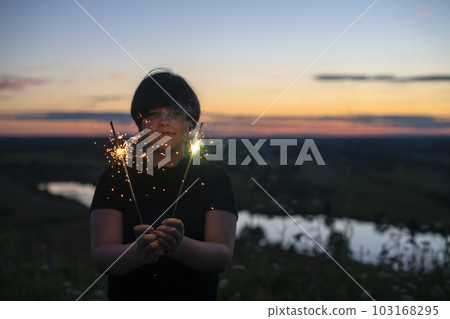 A cute boy holds sparklers in his hands, fireworks in the evening in the summer at sunset. Festive mood A cute boy holds sparklers in his hands, fireworks in the evening in the summer at sunset. Festive mood 103168295