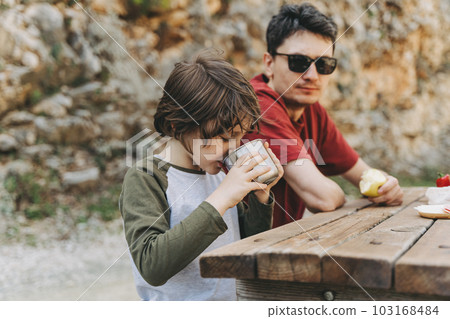 Close-up view of father and his school boy son on a family picnic in the mountains. Child kid and his dad taking a rest and enjoying a picnic while hiking in the mountains 103168484