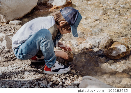 Caucasian school boy throwing rocks into the canyon river. Kid child playing with stones while sitting on a boulder near mountain river bank 103168485