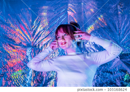 Relaxed smiling woman in white clothes and headphones dancing in neon light. Music lover. White dress code party. Silent disco. DJ girl playing, enjoy moment on shiny background. Selective focus. 103168624
