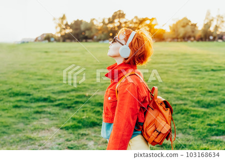 Stylish woman in bright clothes wearing wireless headphones and listening to music while walking on sunset in park. Breathe, close eyes and enjoy the moment. Simple pleasures and personal fulfillment. 103168634