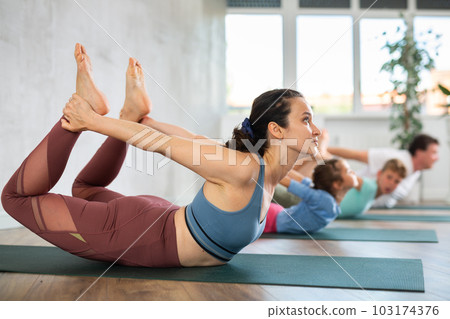 Willing middle-aged woman practicing bow pose of yoga with her family Willing middle-aged woman practicing bow pose of yoga with her family 103174376