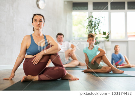 Family of four practicing yoga in fitness studio, posing in pose Gomukhasana 103174452