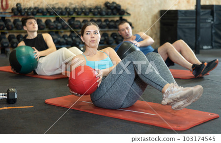 Strong young woman performing exercise for abs core with medicine ball on yoga mat during group workout in CrossFit gym center 103174545