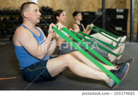 Active middle-aged man wearing sport clothes using resistance band during group workout in gym 103174683