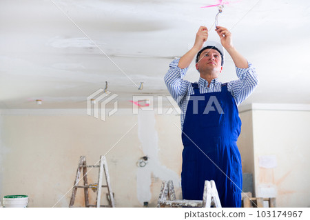 Electrician in blue overalls makes electrical wiring on the ceiling in room being renovated 103174967