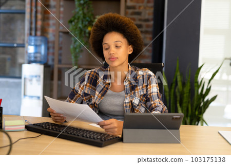 Happy biracial casual businesswoman sitting at desk and reading document 103175138