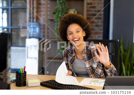 Portrait of happy biracial casual businesswoman making video call at desk, smiling and waving 103175139