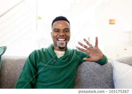 Portrait of african american young man smiling and waving hand while relaxing on sofa in living room Portrait of african american young man smiling and waving hand while relaxing on sofa in living room 103175267