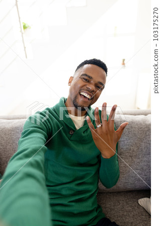 Portrait of happy african american gay man showing wedding ring while sitting on sofa in living room Portrait of happy african american gay man showing wedding ring while sitting on sofa in living room 103175270