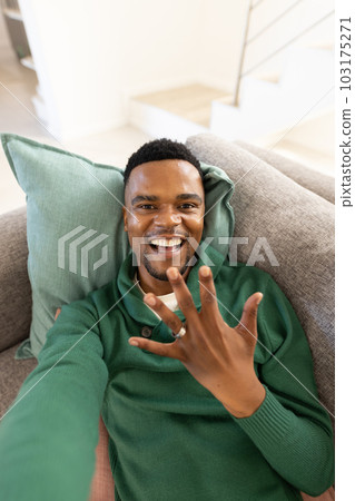 Portrait of happy african american gay man showing wedding ring while lying on sofa in living room 103175271