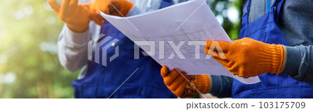 New project. Close up shot of two builders standing outdoors with an open blueprint discussing the cottage construction New project. Close up shot of two builders standing outdoors with an open blueprint discussing the cottage construction 103175709
