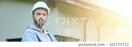 Portrait of young bearded male engineer in hard hat looking at camera, posing outdoors while working on cottage construction Portrait of young bearded male engineer in hard hat looking at camera, posing outdoors while working on cottage construction 103175711
