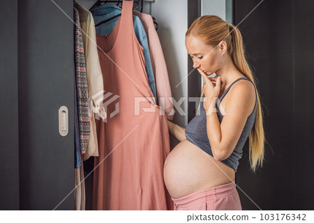 A pregnant woman has nothing to wear. A pregnant woman stands in front of a closet with clothes and does not know what to wear because the clothes do not fit on her 103176342