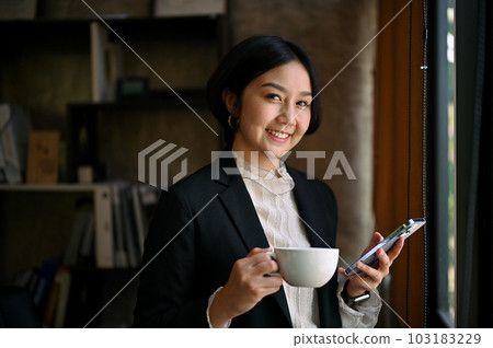 Confident Asian businesswoman standing in her modern office with a coffee cup Confident Asian businesswoman standing in her modern office with a coffee cup 103183229