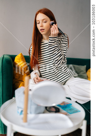 Vertical shot of injured young woman with broken right arm wrapped in white gypsum bandage applying cosmetic on face with professional makeup brush, looking to mirror sitting on sofa at home. Vertical shot of injured young woman with broken right arm wrapped in white gypsum bandage applying cosmetic on face with professional makeup brush, looking to mirror sitting on sofa at home. 103186003