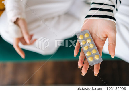 Close-up top view of unrecognizable young woman with broken right hand wrapped in gypsum bandage holding blister packs of pills sitting on couch. Concept of social security and health insurance. Close-up top view of unrecognizable young woman with broken right hand wrapped in gypsum bandage holding blister packs of pills sitting on couch. Concept of social security and health insurance. 103186008
