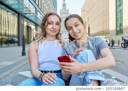 Two smiling female students with smartphone, modern urban style, road, buildings background 103188667