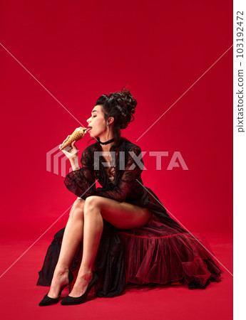 Portrait of young beautiful girl in elegant black dress sitting, eating ice cream against red background 103192472